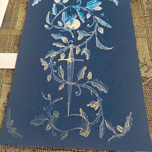 May include: A gold foil print on black paper featuring a stylized illustration of a sword surrounded by an oak tree with acorns and leaves. The moon is visible in the background.