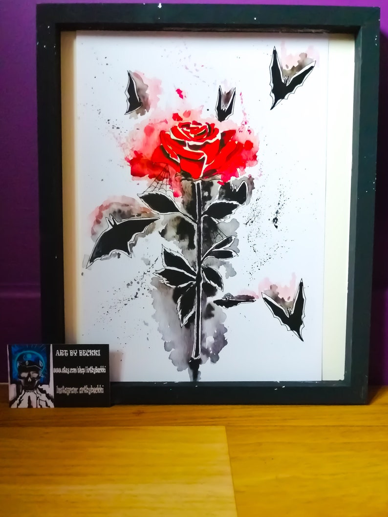 Puede incluir: Una pintura de acuarela enmarcada de una rosa roja con murci&eacute;lagos negros volando a su alrededor. La rosa est&aacute; pintada en un estilo realista con mucho detalle. Los murci&eacute;lagos est&aacute;n pintados de una manera m&aacute;s estilizada. El fondo es una lavada de acuarela azul claro. La pintura est&aacute; firmada "Art by Beckri" en la esquina inferior izquierda.