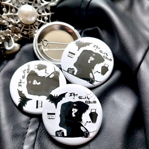 May include: Three white round buttons with black and white illustrations of a person with long black hair and a bat. The text on the buttons reads "The Goth Club".