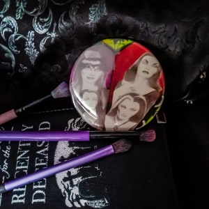 May include: A round, colorful compact mirror with a design of four women's faces. The mirror is on top of a black book with the text "For the Recently Deceased" printed on the cover.