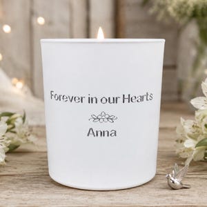 May include: A white candle with the inscription "Forever in our Hearts" and the name "Anna". A small silver bird ornament is placed near the candle, with white flowers in the background.