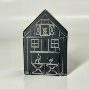 Può includere: Fienile in legno nero con una silhouette bianca di un cavallo e una gallina all'interno. Il fienile ha due finestre e una porta con un design a griglia incrociata.