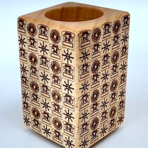 May include: A square wooden pencil holder with a repeating pattern of robot faces and snowflakes. The holder is made of light wood and has a natural finish.