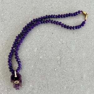 May include: A purple beaded necklace with a Hamsa hand pendant. The pendant is a carved purple stone with a gold-colored accent. The necklace is made of small, faceted purple beads and has a gold clasp.