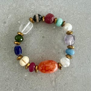 以下が含まれることがあります： 赤、オレンジ、青、緑、白の色合いの様々な宝石ビーズが特徴のカラフルなビーズブレスレット。 ブレスレットには、ゴールドカラーのスペーサーと透明な長方形のビーズが含まれています。 周囲は約18cmです。