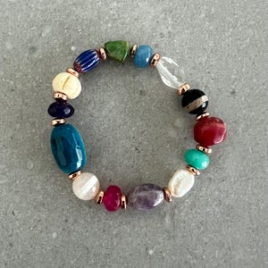 May include: A colorful beaded bracelet featuring a variety of gemstone beads in shades of blue, green, red, white, and purple. The beads are connected with rose gold-colored metal accents. The bracelet is a stylish accessory.