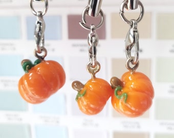Può includere: Tre ciondoli a forma di zucca arancione con steli e foglie verdi appesi a catene d'argento. I ciondoli sono attaccati a cordini neri, gialli e arancioni.