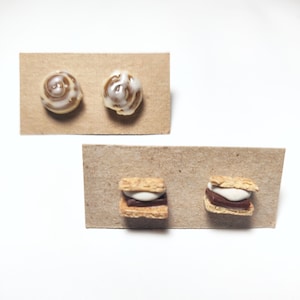 May include: Two pairs of polymer clay earrings. The first pair is shaped like cinnamon rolls with brown and white icing. The second pair is shaped like s'mores with graham crackers, chocolate, and marshmallow.