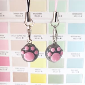 May include: Two grey cat paw charms with pink paw pads, attached to silver keychains. The charms are hanging from a white and light blue background.