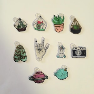 May include: Ten colorful charms with various designs, including a plant in a glass dome, a unicorn, a camera, a hand making the rock and roll sign, a planet, a narwhal, and a leaf. The charms are made of clear plastic and are perfect for adding a touch of whimsy to jewelry or crafts.