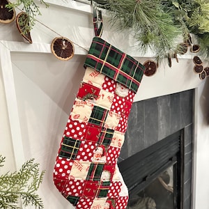 Può includere: Un calzino di Natale in patchwork rosso e verde con un polsino a quadri. Il calzino è decorato con pois bianchi, volti di Babbo Natale e la parola "Motel".