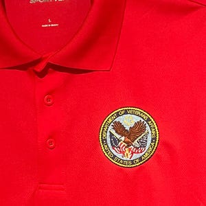 May include: A bright red polo shirt with a collar and three buttons. The shirt features a circular emblem with an eagle and the text "DEPARTMENT OF VETERANS AFFAIRS UNITED STATES OF AMERICA". The brand name "SPORT-TEK" is visible on the collar.