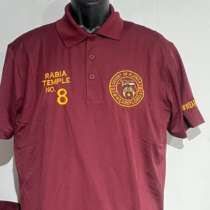 May include: A maroon polo shirt with a collar and three buttons. The shirt features embroidered text and a circular patch on the chest that reads "Desert of Florida". The left chest has the text "RABIA TEMPLE NO. 8". The sleeve has the text "#WEARFLORIDA".