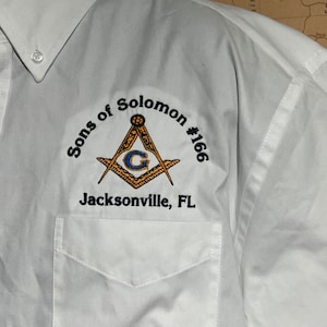May include: White button-down shirt with a pocket and embroidered emblem. The emblem features a compass and square design with the text "Sons of Solomon #166 Jacksonville, FL".