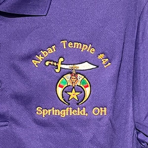 May include: Purple polo shirt with embroidered text and emblem. The text reads "Akbar Temple #41" and "Springfield, OH" in gold thread. The emblem features a sword, crescent moon, star, and other symbols.