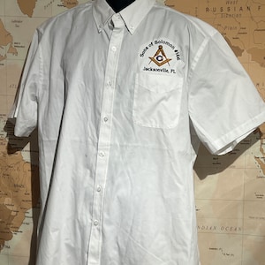 May include: White short-sleeved button-down shirt with a pocket on the left side. The pocket has an embroidered patch that says "Sons of Solomon #166 Jacksonville, FL" and features a compass and square.