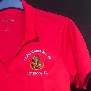 May include: A red polo shirt with the text "Malta Court No. 99 Orlando, FL" embroidered in white and gold. The shirt has short sleeves and a collared neckline. The brand name "SPORT-TEK" is visible on the upper left.