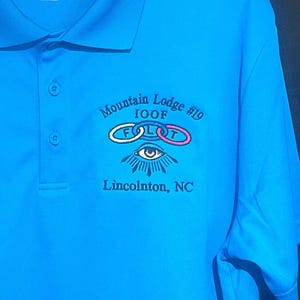 May include: Bright blue polo shirt with the text "Mountain Lodge #19 IOOF" and an emblem featuring interlocking rings and an eye. The shirt is a size XL and has the Sport-Tek brand logo.