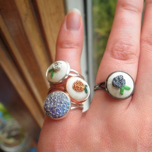 May include: A hand wearing four rings with embroidered flowers. The rings are silver and have different coloured flowers. The flowers are white, orange, blue, and black.