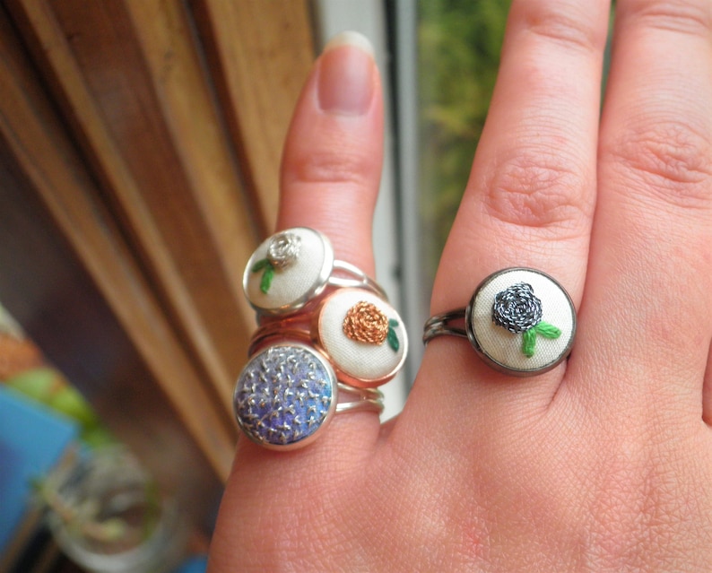 May include: A hand wearing four rings with embroidered flowers. The rings are silver and have different coloured flowers. The flowers are white, orange, blue, and black.