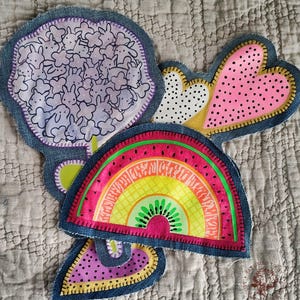 Könnte beinhalten: Eine Sammlung farbenfroher Stoffaufnäher mit Jeanskanten. Zu den Designs gehören ein rosa Herz mit schwarzen Punkten, ein Regenbogen mit Kiwi-Mitte und eine lila Form mit Hasenillustrationen. Die Aufnäher befinden sich auf einem strukturierten, neutralen Hintergrund.