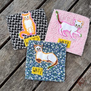 Op de afbeelding: Drie vierkante stoffen patches met kattenontwerpen. Eén patch heeft een oranje kat op een zwart-wit patroonachtergrond. Een andere heeft een witte kat op een roze achtergrond. De derde patch heeft een wit-bruine kat op een blauwe bloemenachtergrond. Elke patch heeft een geel label met het woord "WAT?"