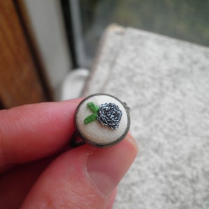 May include: A silver ring with a white fabric base and a small embroidered black flower with green leaves.