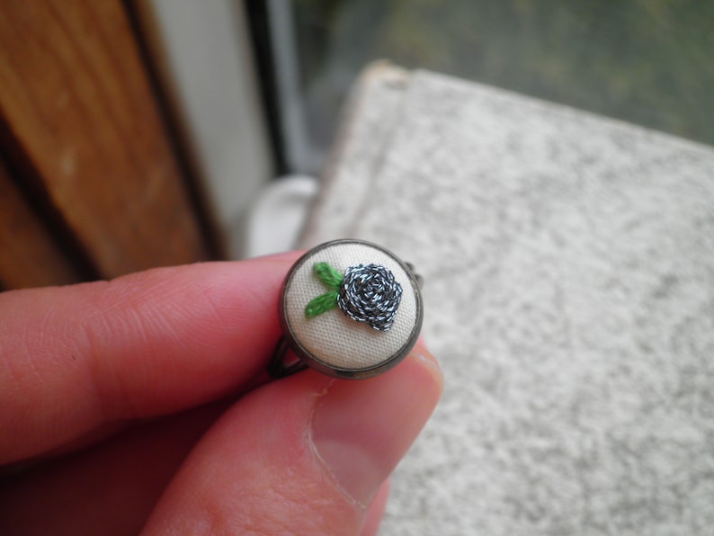May include: A silver ring with a white fabric base and a small embroidered black flower with green leaves.