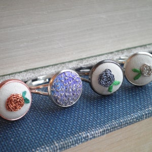 May include: Five silver rings with white fabric centres featuring embroidered floral designs. The designs include a purple flower, a black flower, a white flower, and two orange flowers.