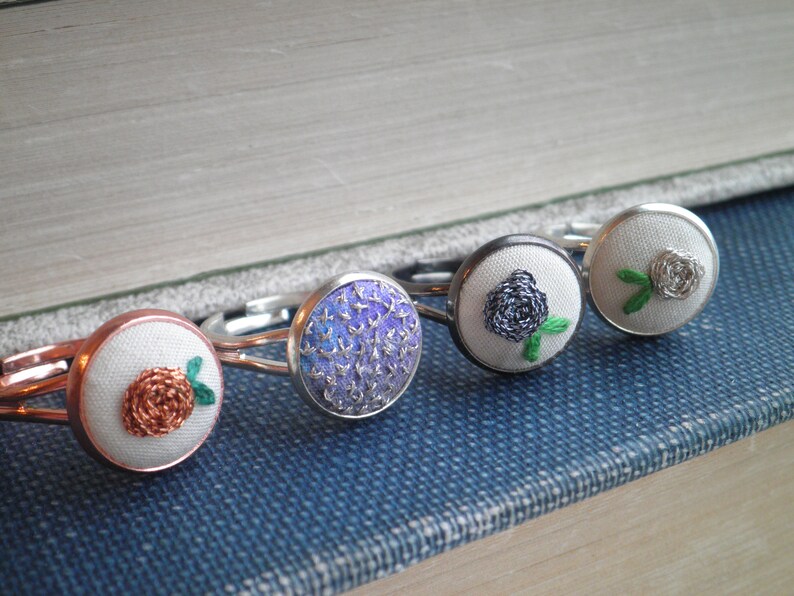 May include: Five silver rings with white fabric centres featuring embroidered floral designs. The designs include a purple flower, a black flower, a white flower, and two orange flowers.