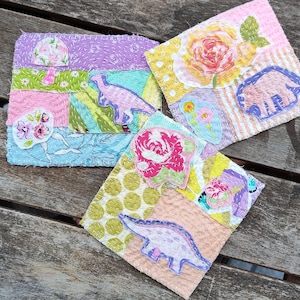 Op de afbeelding: Drie handgemaakte stoffen onderzetters met patchwork ontwerpen. Elke onderzetter heeft bloemen- en dinosaurus applicaties in pastelkleuren. De onderzetters zijn gestikt met witte draad en liggen op een verweerd houten oppervlak.