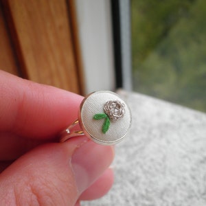 May include: A silver ring with a white fabric base and a white embroidered rose with green leaves.