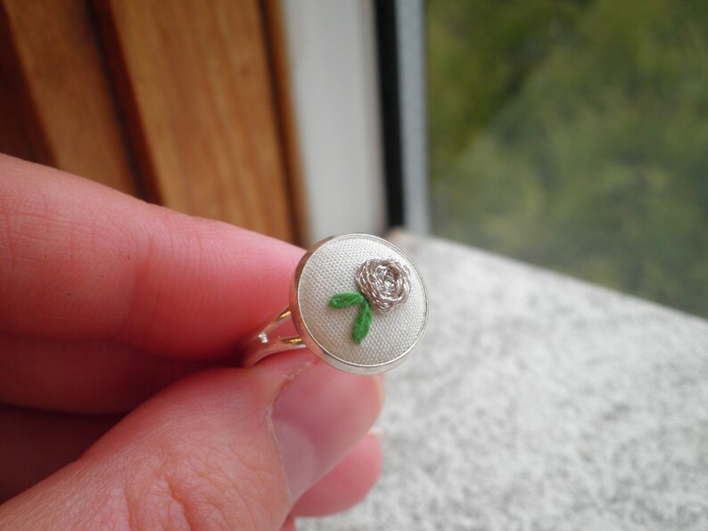 May include: A silver ring with a white fabric base and a white embroidered rose with green leaves.