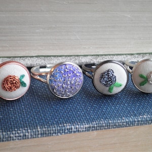 May include: Four silver rings with different embroidered fabric designs. The rings feature a rose, a blue flower, a black flower, and a white flower.