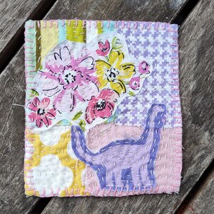 Op de afbeelding: Een handgemaakt textielkunstwerk met een dinosaurus- en bloemenontwerp. Het kunstwerk toont een paars dinosaurus silhouet en een boeket roze en gele bloemen. De achtergrond bevat verschillende stoffen met patronen, allemaal aan elkaar gestikt met zichtbare draad.