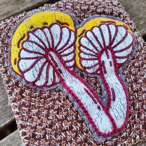 Pode incluir: Um close-up de um remendo bordado feito à mão com dois cogumelos brancos com detalhes vermelhos sobre um fundo de tecido marrom com um padrão floral repetitivo.