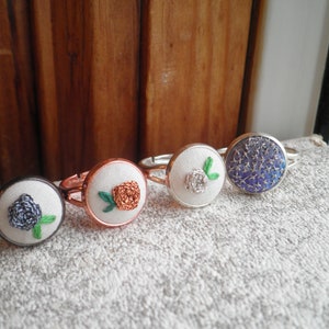 May include: Four silver rings with different embroidered designs. The rings feature a white flower, a blue flower, a brown flower, and a blue and silver design.