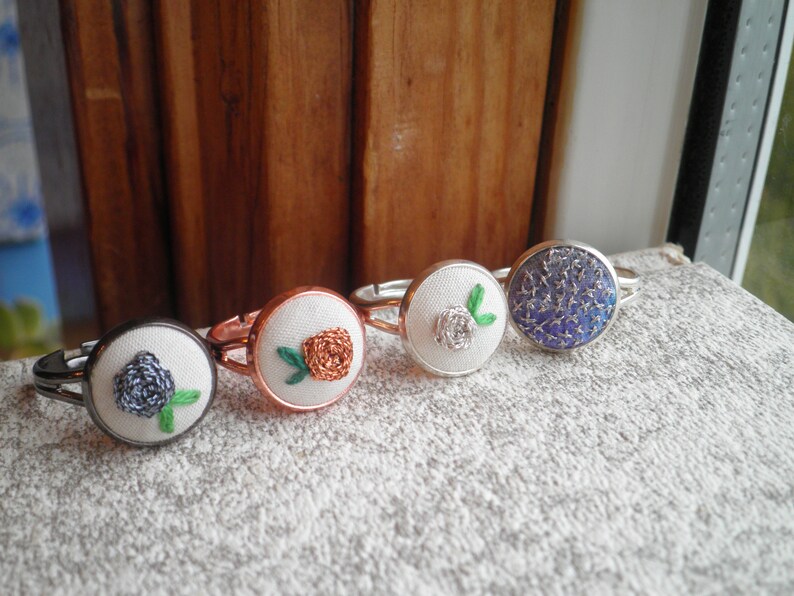 May include: Four silver rings with different embroidered designs. The rings feature a white flower, a blue flower, a brown flower, and a blue and silver design.