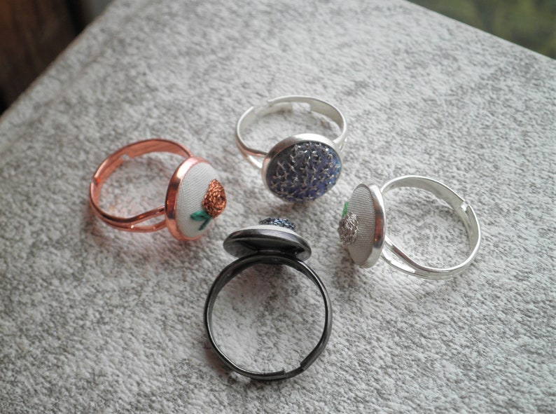 May include: Four rings with different coloured and patterned buttons. The rings are made of silver, copper, and black metal. The buttons are round and have a variety of designs.