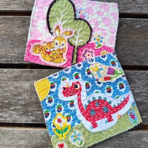 May include: Two handmade fabric squares with colorful embroidered designs. The first square features a pink background with a green tree, a brown squirrel, and a brown rabbit. The second square features a blue background with a red dinosaur, yellow flowers, and green grass.