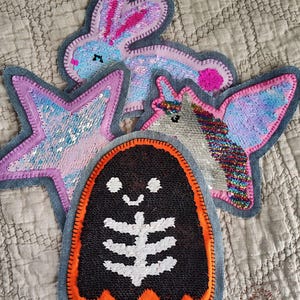 Op de afbeelding: Een verzameling kleurrijke pailletten patches in verschillende vormen: een konijn, een ster, een spook en een gevleugeld wezen. Elke patch is omrand met denim en heeft heldere kleuren zoals roze, blauw en oranje. De patches zijn decoratief.