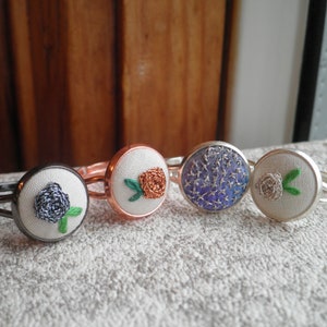 May include: Four silver rings with different embroidered fabric flowers. The flowers are a blue rose, an orange rose, a blue and silver floral pattern, and a white rose.
