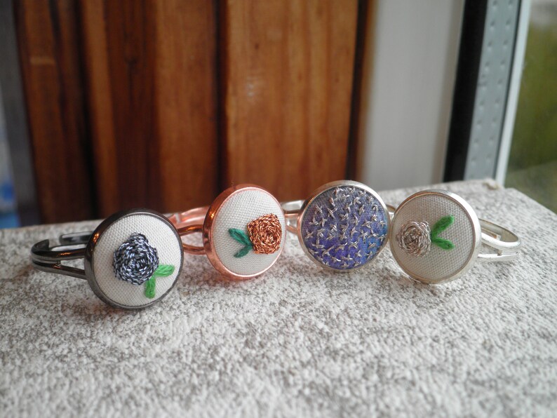 May include: Four silver rings with different embroidered fabric flowers. The flowers are a blue rose, an orange rose, a blue and silver floral pattern, and a white rose.
