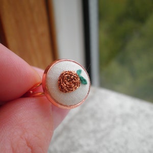 May include: A copper ring with a white fabric centre featuring a small embroidered flower with green leaves.