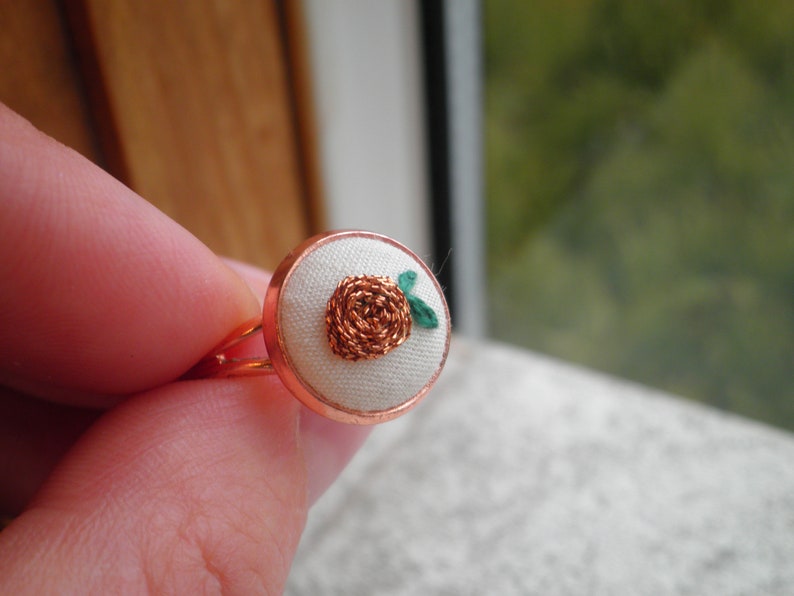 May include: A copper ring with a white fabric centre featuring a small embroidered flower with green leaves.
