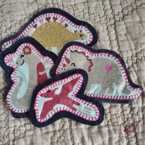 May include: Five dinosaur-shaped patches with glittery accents. The patches feature various colors, including gold, silver, and red, with denim blue borders and pink stitching. Each dinosaur has a unique design, including hearts and flowers.