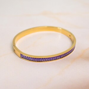May include: A gold-colored bangle bracelet with a row of small, rectangular purple gemstones. The bracelet is circular and has a simple, elegant design, suitable for everyday wear or special occasions.