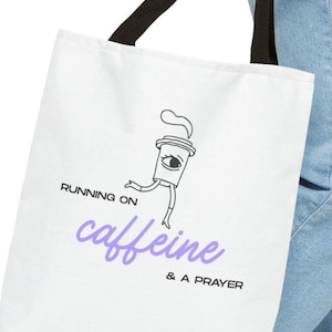 May include: White tote bag with black handles featuring a cartoon coffee cup with an eye, running. The text reads "RUNNING ON caffeine & A PRAYER" in black and purple lettering. The bag is made of a sturdy fabric.