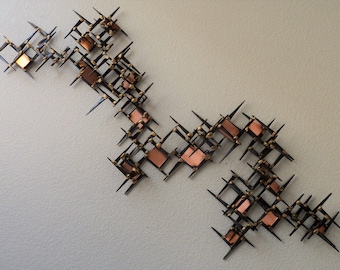 May include: Abstract wall art made of metal with a geometric design. The piece features a series of interconnected squares and triangles, with a copper finish. The artwork is mounted on a white wall.