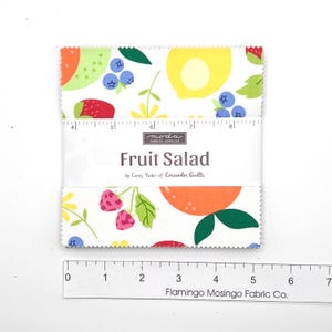 May include: A square stack of fabric squares featuring a white background with colorful fruit illustrations including strawberries, blueberries, oranges, and lemons. A paper band reads "Fruit Salad by Corey Yoder of Coriander Quills" with a 5-inch ruler.